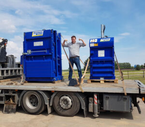 A man stands on a wheeled lorry platform in between two large Strautmann balers. He is stood in a strong-man pose, smiling.