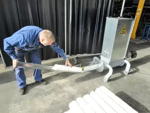 A man stands bent over as he pulls compacted Styrofoam tubes from the Styropactor Nano. The Styropactor Nano has four curved legs and a sticker of two eyes on the front of its metal cuboid shell.